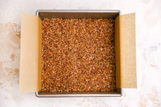 A square baking pan lined with parchment paper. A date crust is pressed into the bottom.
