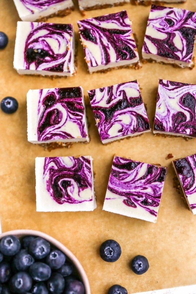 Overhead photo of a sheet of parchment paper with vegan blueberry cheesecake squares scattered around.