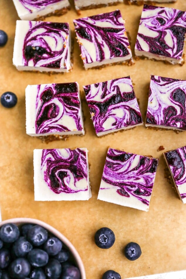 Overhead photo of a sheet of parchment paper with vegan blueberry cheesecake squares scattered around.