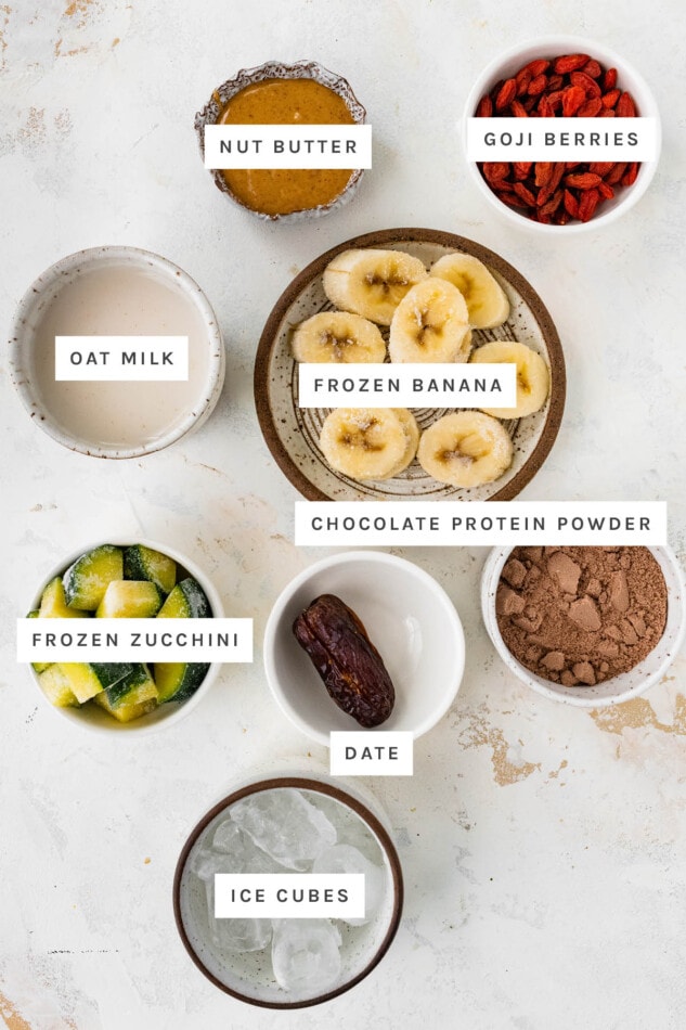 Ingredients measured out to make a Butterfinger Smoothie: nut butter, goji berries, oat milk, frozen banana, chocolate protein powder, frozen zucchini, date and ice cubes.