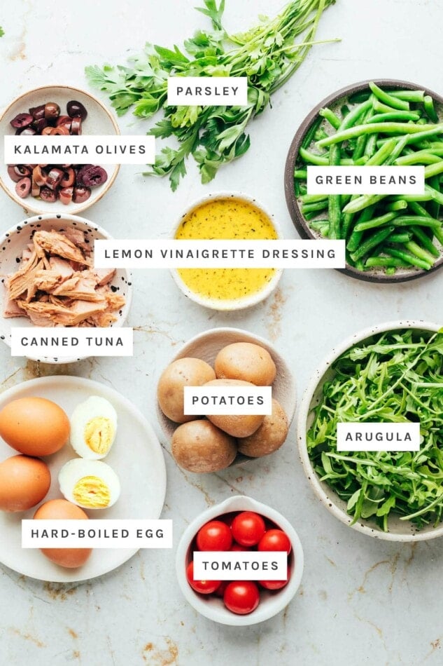 Ingredients measured out to make nicoise salad: parsley, kalamata olives, green beans, lemon vinaigrette dressing, canned tuna, potatoes, arugula, hard-boiled egg and tomatoes.