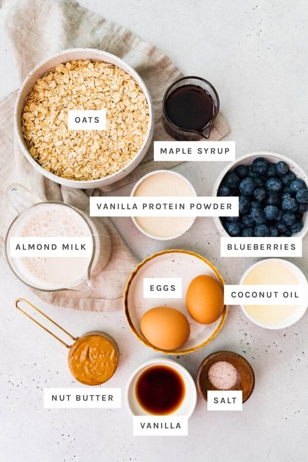 Ingredients measured out to make protein baked oatmeal: oats, maple syrup, vanilla protein powder, blueberries, almond milk, eggs, coconut oil, nut butter, vanilla and salt.