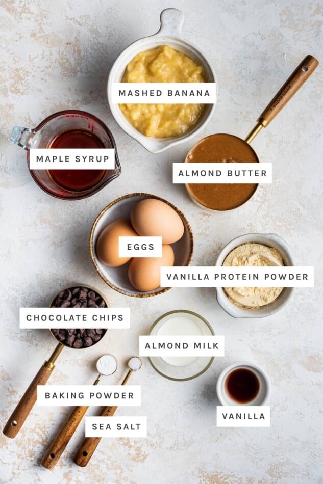 Ingredients measured out to make Protein Muffins: mashed banana, maple syrup, almond butter, eggs, vanilla protein powder, chocolate chips, almond milk, baking powder, sea salt and vanilla.