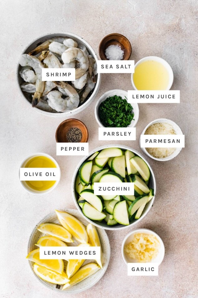 Ingredients measured out to make Shrimp Scampi Sheet Pan Meal: shrimp, sea salt, lemon juice, parsley, parmesan, pepper, olive oil, zucchini, lemon wedges and garlic.