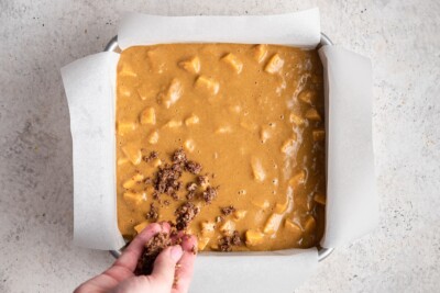 Cake batter in a square baking pan lined with parchment paper. A hand is drizzling the topping over top of the batter.