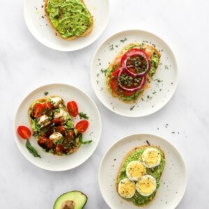 Four styles of avocado toast on four plates with an open, halved avocado.