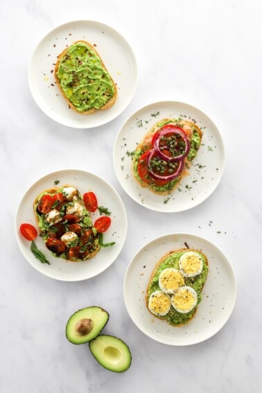 Four styles of avocado toast on four plates with an open, halved avocado.