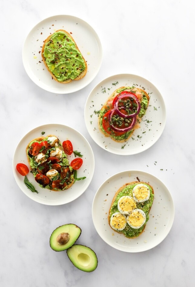Four styles of avocado toast on four plates with an open, halved avocado.