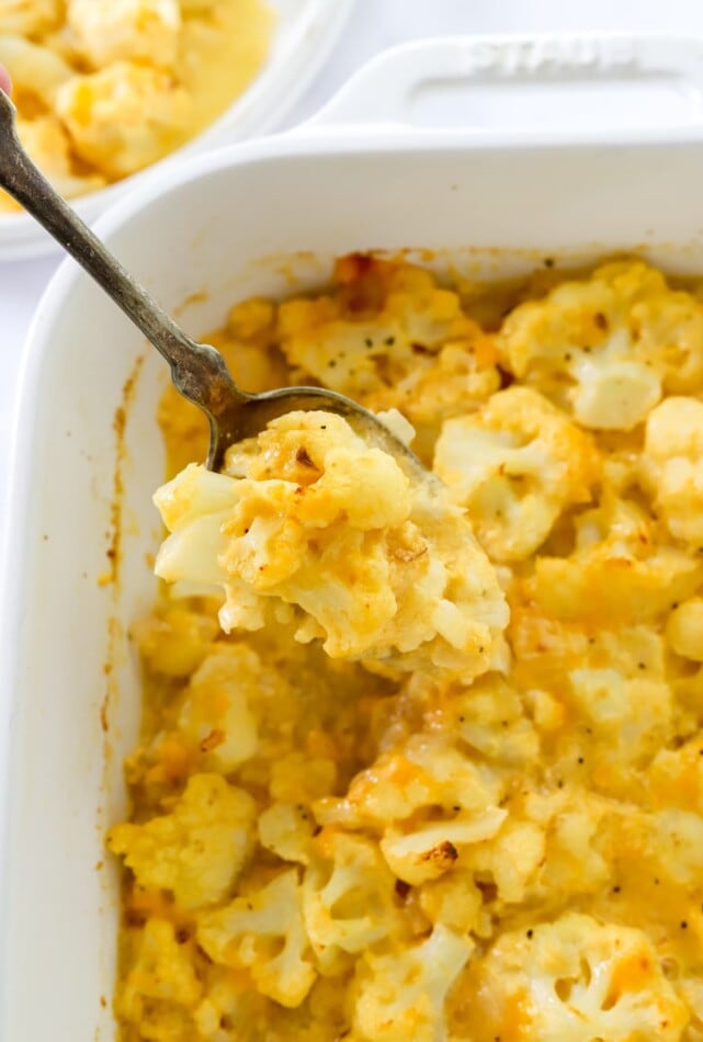 A spoon lifting up a serving of baked cauliflower mac and cheese out of a baking dish.