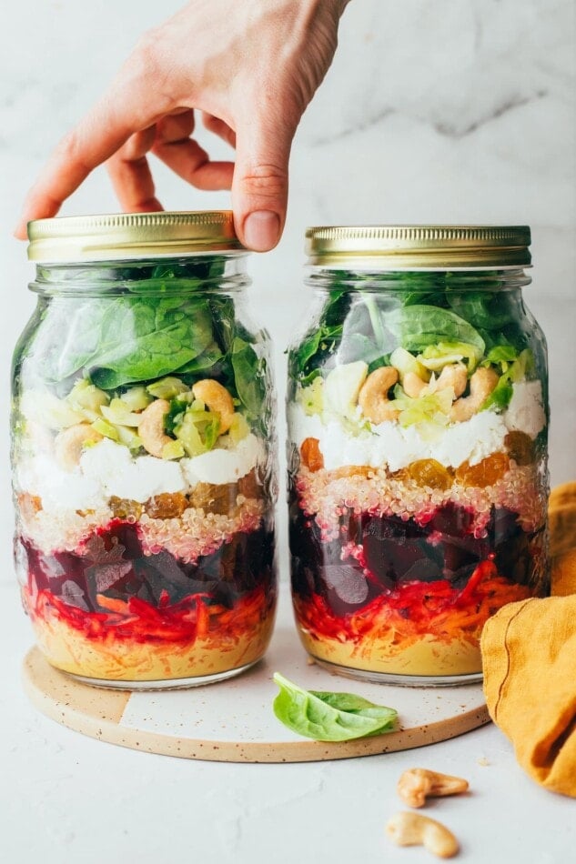 Two jars side by side containing beet power salad ingredients. One jar has a lid and the other jar has a hand twisting a lid on.