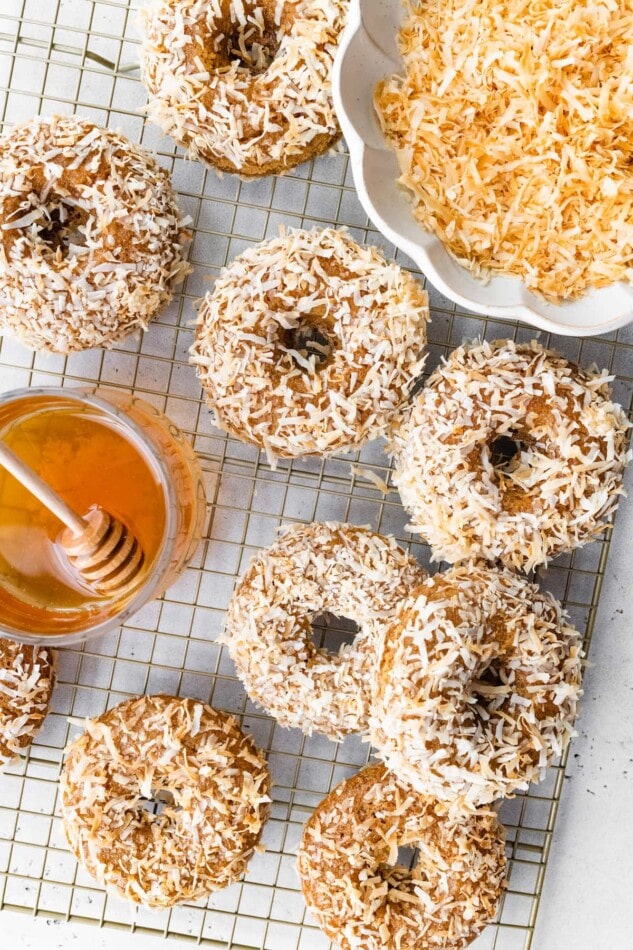 Overhead view of coconut crunch donuts on a wire rack with a bowl of toasted coconut flakes and a bowl of honey.