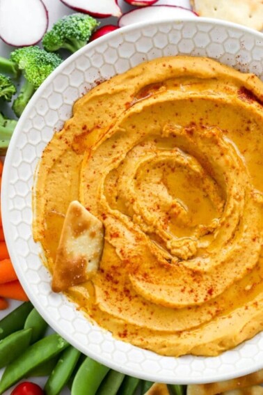 Close up of sweet potato hummus in a bowl. A pita chip rests in the hummus.
