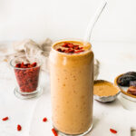 A butterfinger smoothie in a glass with a straw topped with goji berries.