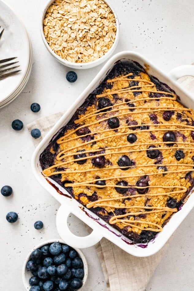 Protein baked oatmeal in a baking dish with bowls of blueberries and oats around it.