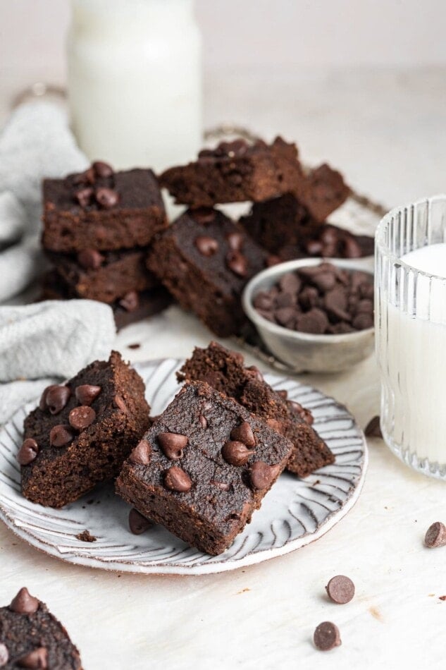 A plate with three brownies with a glass of milk. More brownies are in the background out of focus.