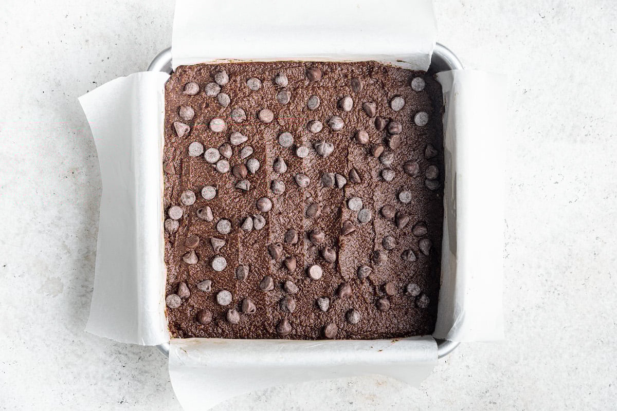 Brownies batter spread across a square baking pan lined in parchment paper. Additional chocolate chips have been decorated across the top of the batter.