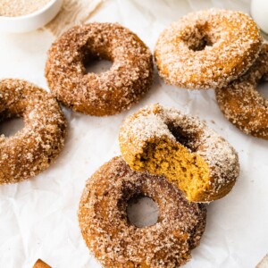Pumpkin spice donuts on a sheet of parchment paper. Two donuts are resting against each other and the top one has a bite missing.