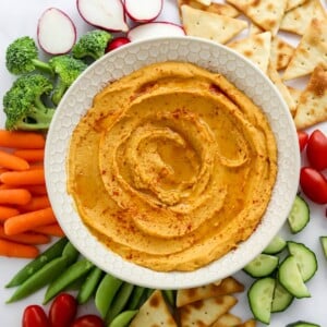 A bowl of sweet potato hummus surrounded by various vegetables and pita chips for dipping.