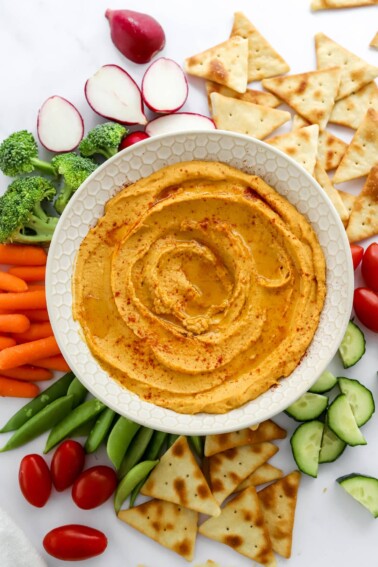 A bowl of sweet potato hummus surrounded by various vegetables and pita chips for dipping.
