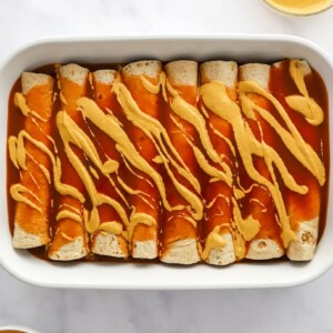 Enchiladas in a row in a baking dish topped with enchilada sauce and vegan queso.