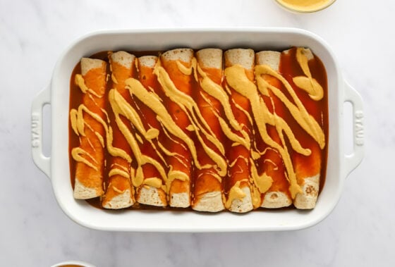 Enchiladas in a row in a baking dish topped with enchilada sauce and vegan queso.