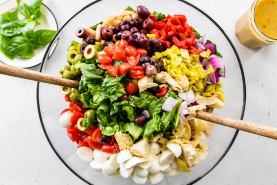 Ingredients for antipasto salad being mixed together with two wooden utensils in a large glass mixing bowl.