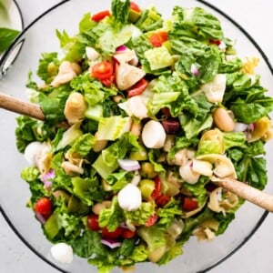 Antipasto salad in a large glass mixing bowl with wooden utensils.