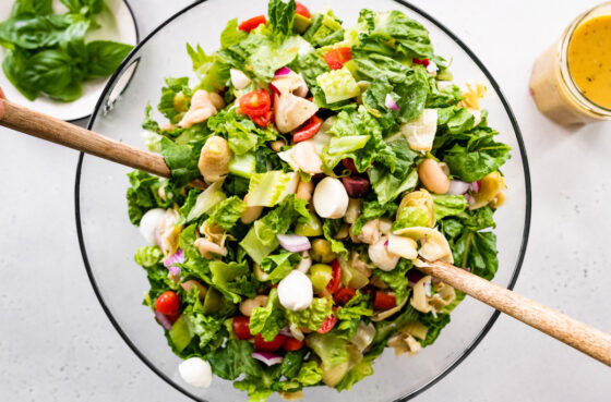 Antipasto salad in a large glass mixing bowl with wooden utensils.