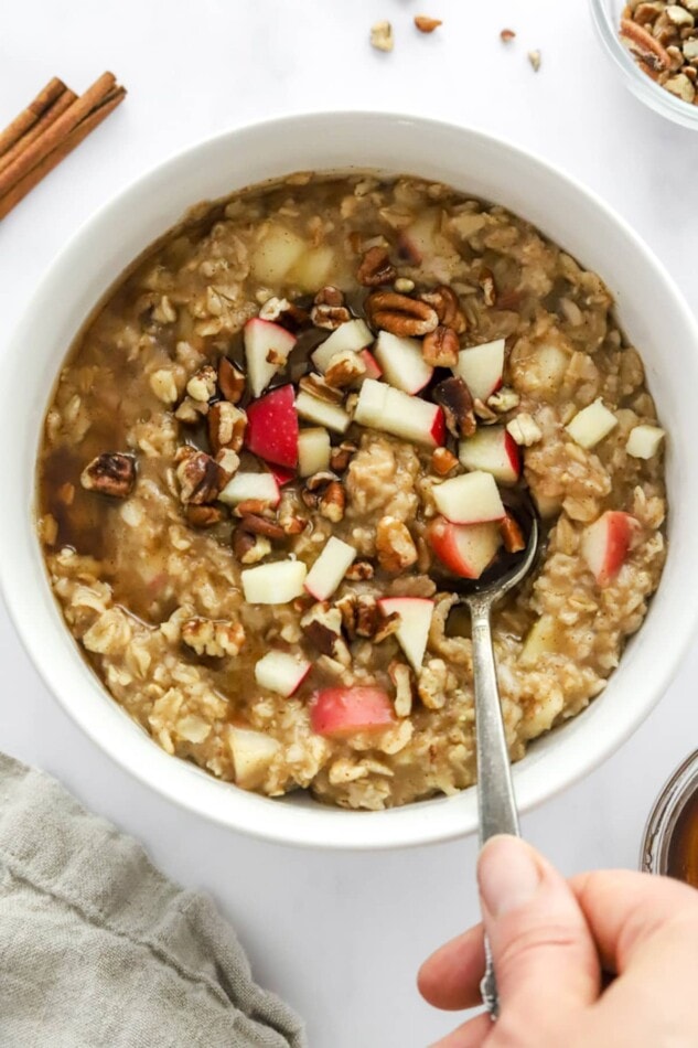 A hand holding a spoon, scooping out a bite of apple cinnamon oatmeal from a bowl.