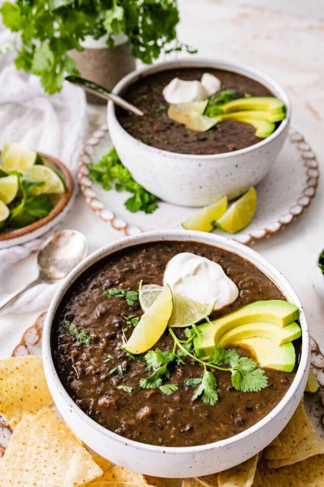 Two bowls of black bean soup topped with avocado, cilantro, lime and vegan sour cream.
