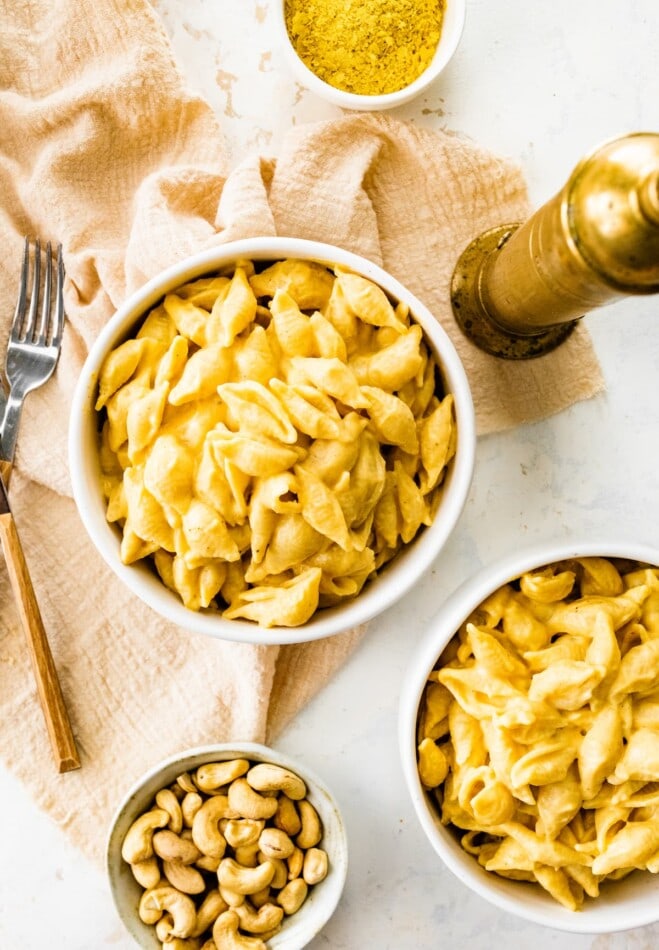 Two bowls of mac and cheese with a cloth napkin, forks, and bowls of cashews and nutritional yeast, plus a pepper grinder.