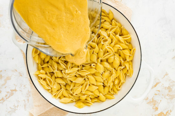 Pouring vegan cheese over cooked noodles in a pot.