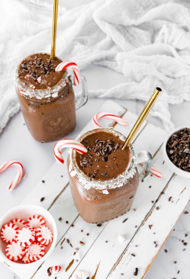 Two glass mugs containing chocolate peppermint smoothies with candy canes and metal straws.