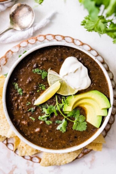 A bowl of black bean soup topped with avocado, cilantro, lime and vegan sour cream served alongside tortilla chips.