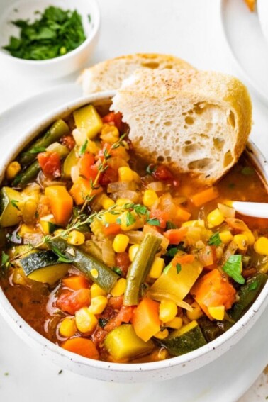 A bowl of vegetable soup with a chunk of bread dipped in the soup.
