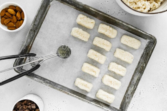 A cookie scoop lays on a parchment lined baking sheet alongside coconut mixture shaped into rectangular bars.