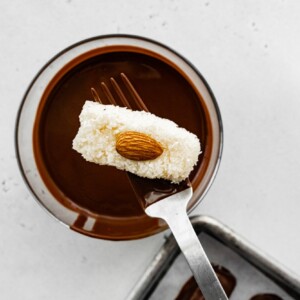 A fork dipping a coconut bar with an almond into a bowl of melted chocolate to coat it.