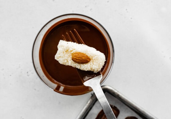 A fork dipping a coconut bar with an almond into a bowl of melted chocolate to coat it.