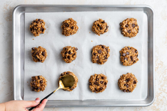 Twelve cookies on a baking sheet, a spoon is being pressed into the cookies to flatten them a bit.