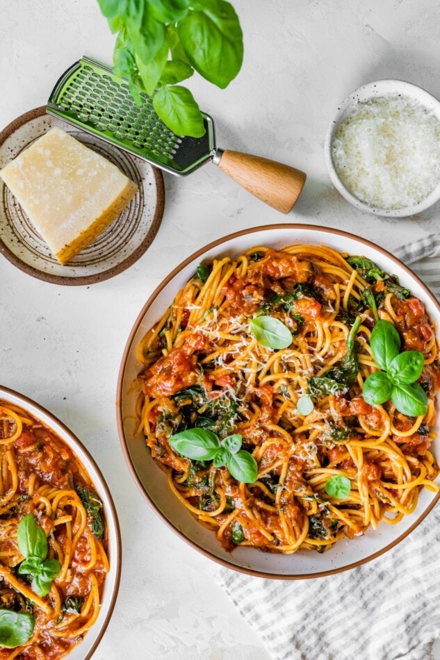 Two bowls of pasta topped with fresh basil leaves. A small plate holds a chunk of parmesan, a hand held cheese grater lays next to it.