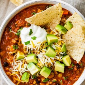 A bowl of taco soup topped with avocado chunks, shredded cheese and sour cream with tortilla chips.
