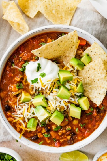 A bowl of taco soup topped with avocado chunks, shredded cheese and sour cream with tortilla chips.