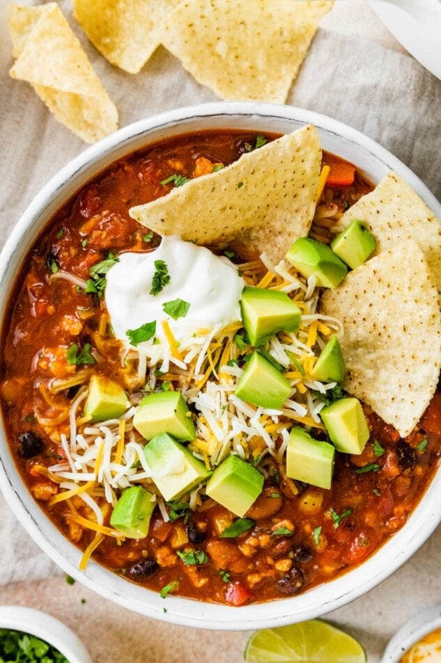 A bowl of taco soup topped with avocado chunks, shredded cheese and sour cream with tortilla chips.