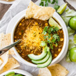 A bowl of vegetarian chili verde served with avocado, lime, cheese, cilantro and a tortilla.
