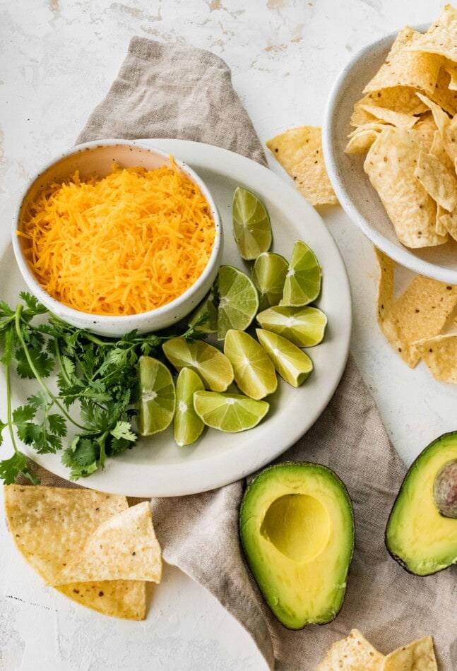 A bowl of shredded cheese with fresh cilantro, lime wedges, avocados and tortilla chips for serving with chili verde.
