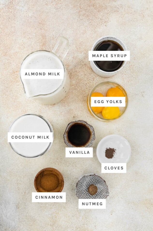 Ingredients measured out to make Dairy-Free Eggnog: almond milk, maple syrup, egg yolks, coconut milk, vanilla, cloves, cinnamon and nutmeg.