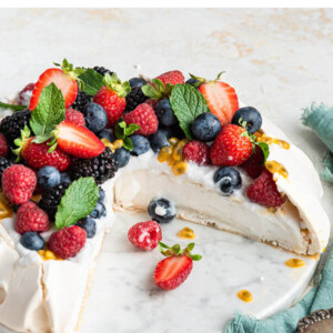 A pavlova topped with coconut whipped cream and fresh fruit with slices missing.