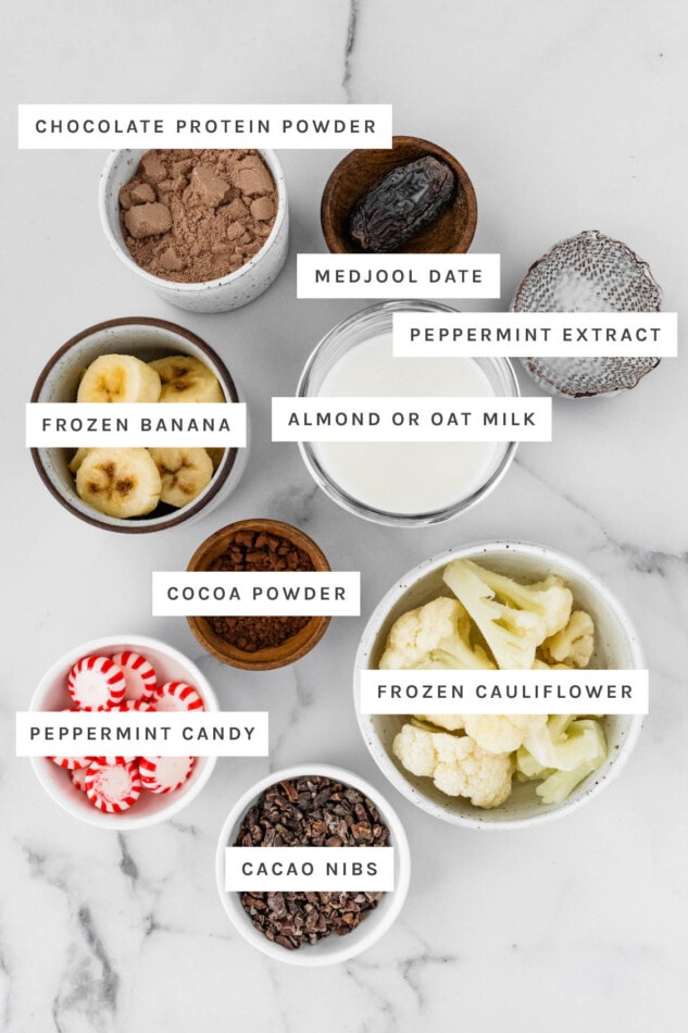 Ingredients measured out to make Chocolate Peppermint Smoothie: chocolate protein powder, medjool date, peppermint extract, frozen banana, almond/oat milk, cocoa powder, frozen cauliflower, peppermint candy and cacao nibs.