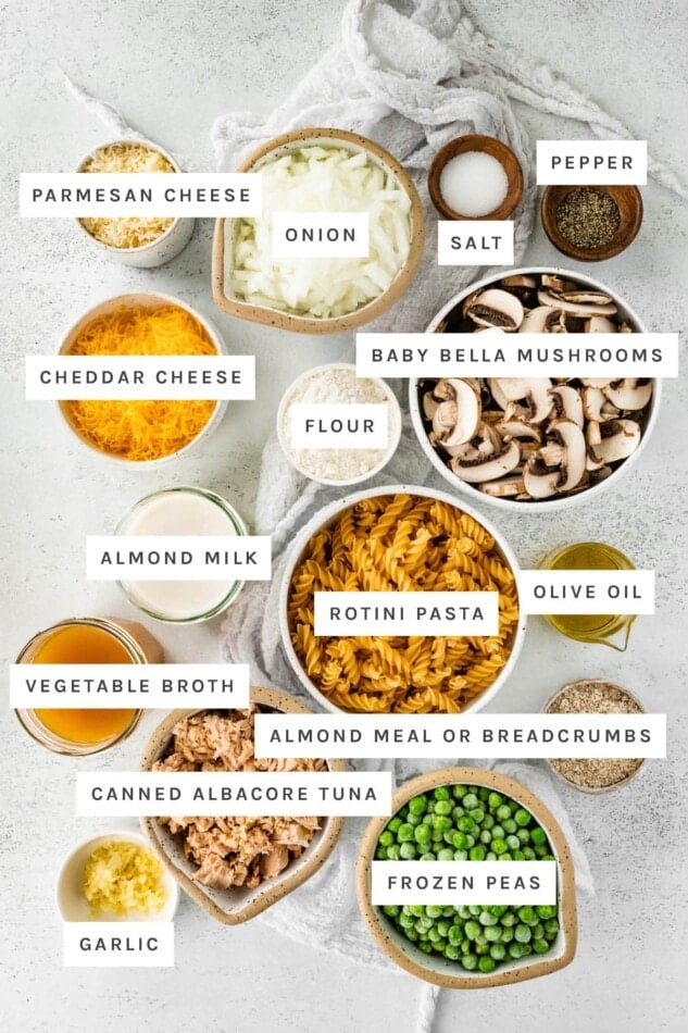 Ingredients measured out to make Healthy Tuna Noodle Casserole: parmesan, onion, salt, pepper, cheddar, flour, baby bella mushrooms, almond milk, rotini pasta, olive oil, vegetable broth, almond meal or breadcrumbs, canned tuna, frozen peas and garlic.