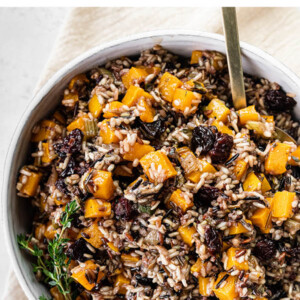Bowl of wild rice stuffing with butternut squash and dried cherries. Garnished with thyme. Serving spoon is in the bowl.